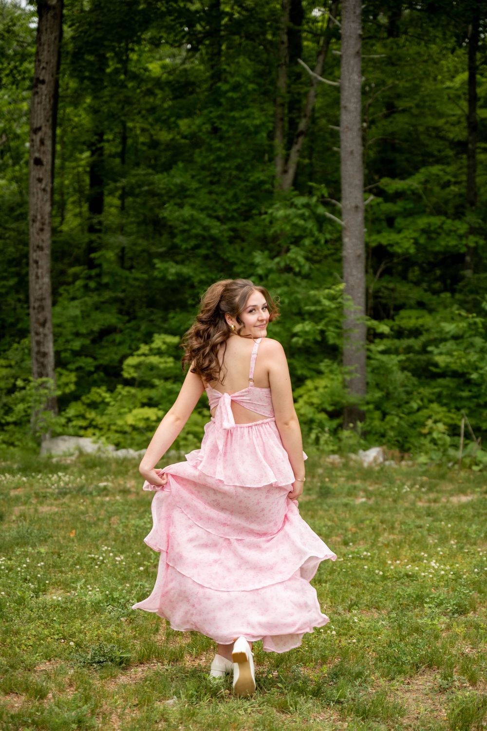 A person in a flowing pink polka dot dress twirls playfully in a sunlit meadow surrounded by tall forest trees.