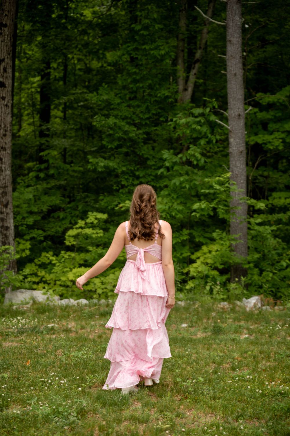 A figure in a flowing pink tiered dress walks through a grassy clearing towards a lush green forest.
