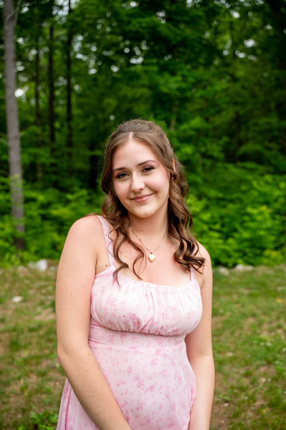 A woman in a pink floral dress poses in an outdoor wooded setting with lush green trees in the background.