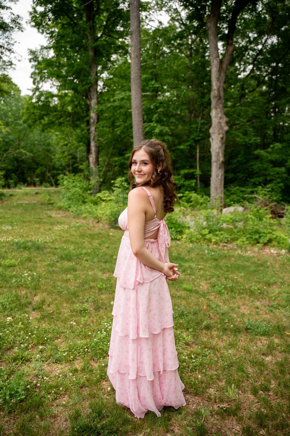 Someone in a flowing pink tiered dress poses playfully in a sunny wooded area.