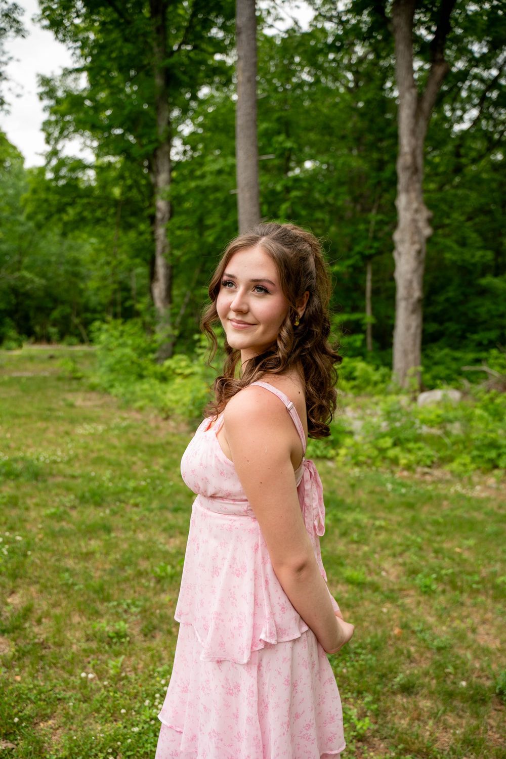 Woman wearing a flowing pink dress stands in a tranquil forest clearing surrounded by tall trees.