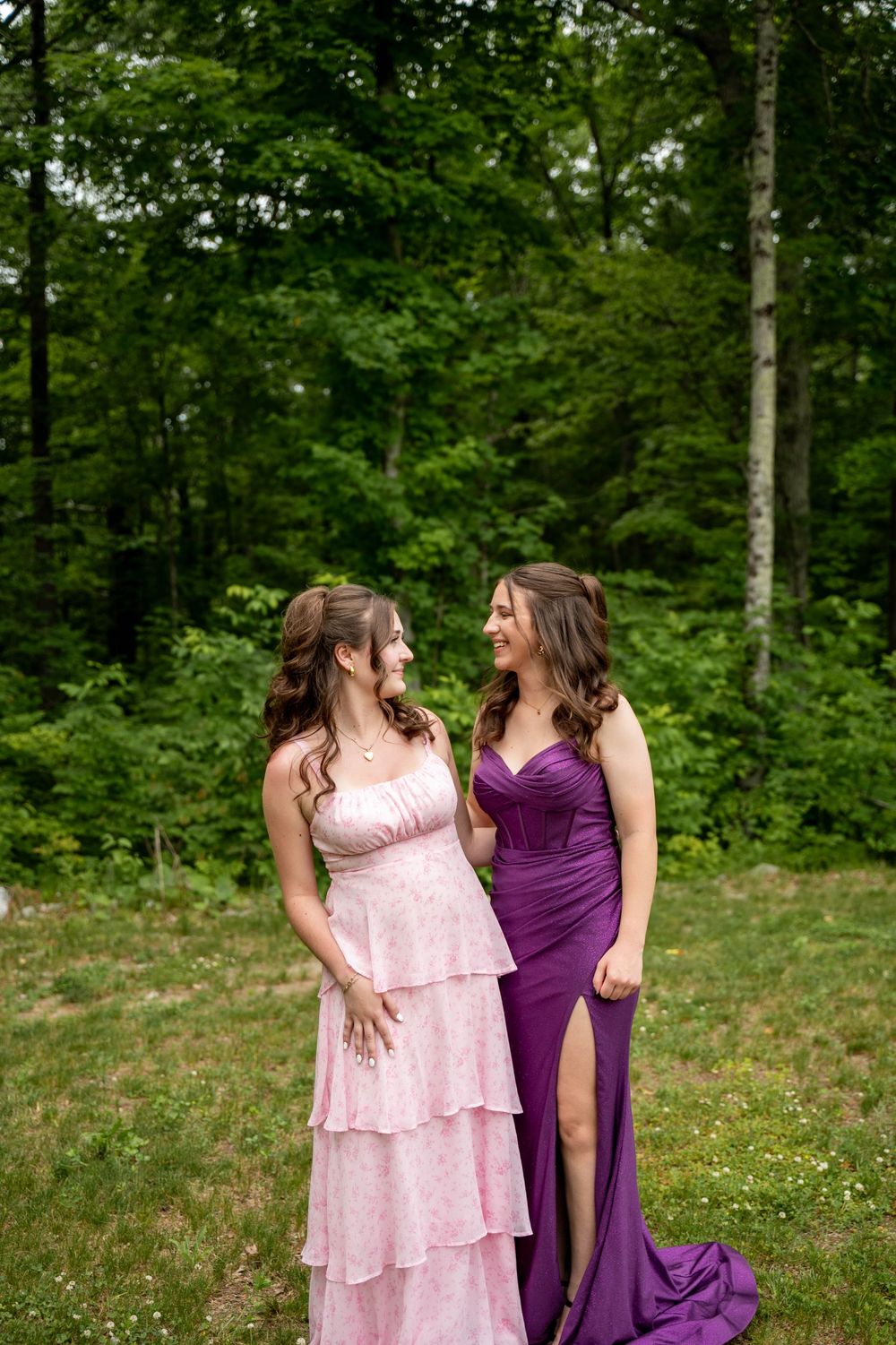 Two bridesmaids share a joyful moment in a forest clearing, wearing pink and purple formal gowns.