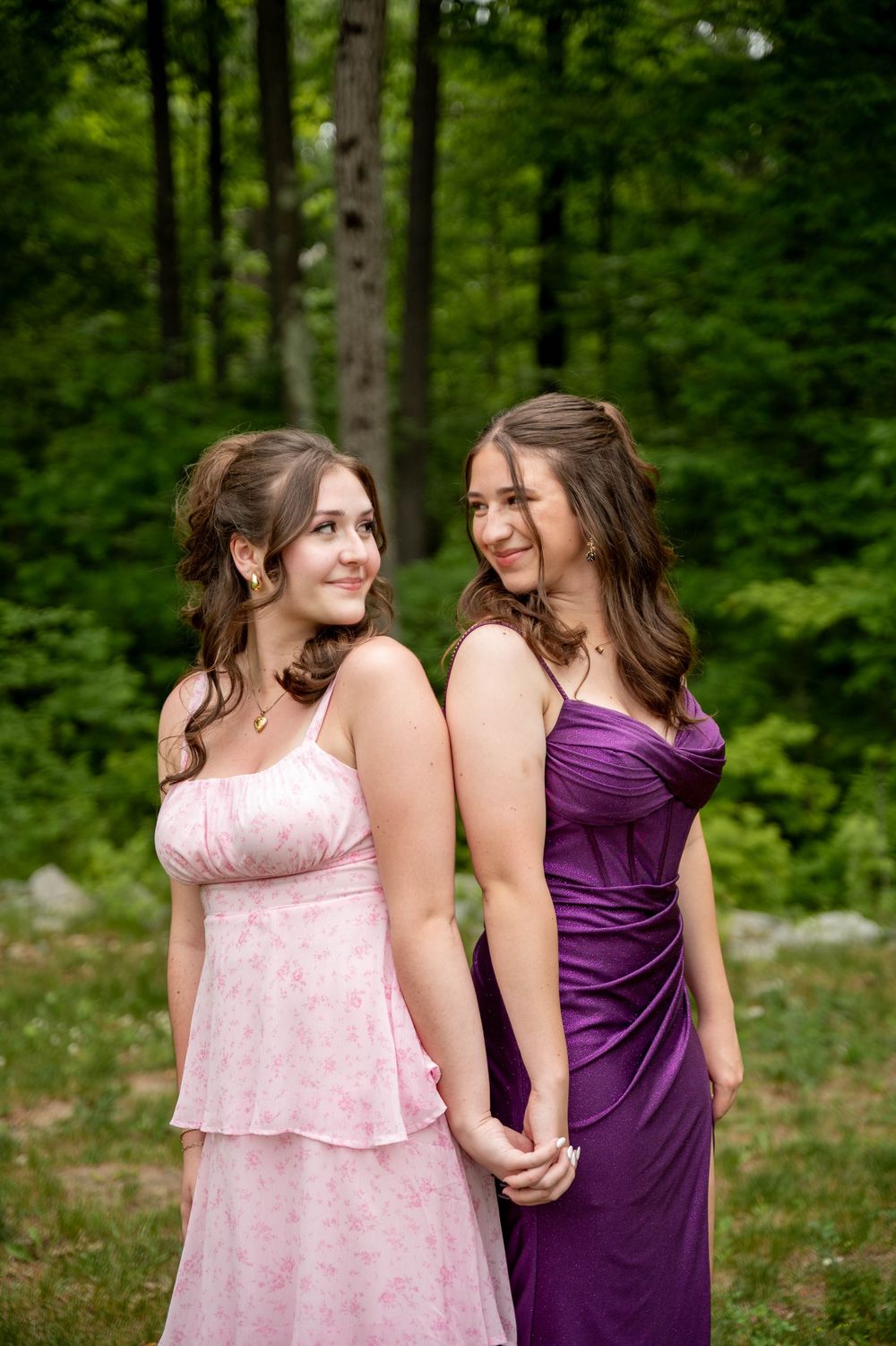 Two friends in pink and purple dresses share a playful moment in a wooded outdoor setting.