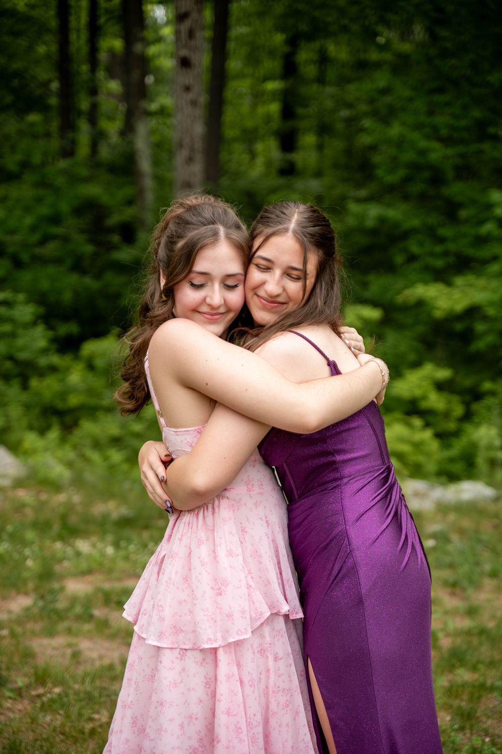 Two people in formal dresses, one pink and one purple, share an embrace outdoors in a wooded setting.