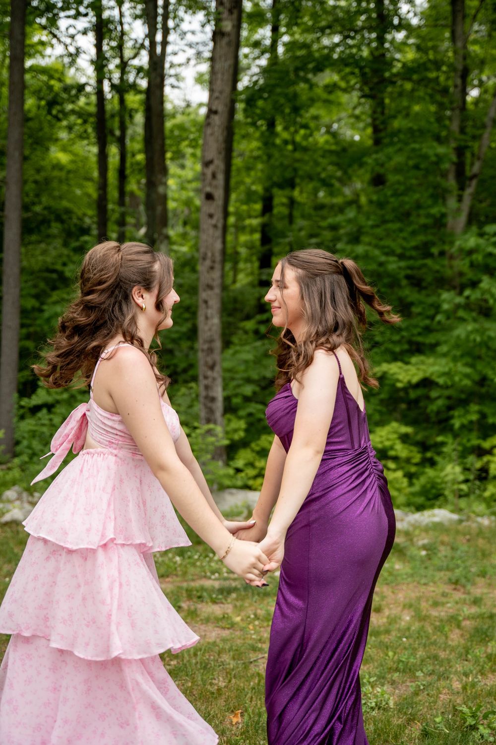 A duo in contrasting pink and purple dresses dance together on grass with trees in the background.