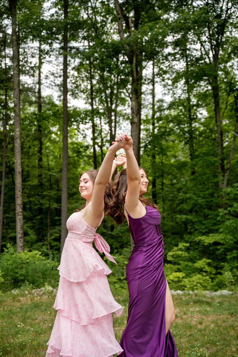 Two friends in pink and purple dresses dance joyfully together in a wooded outdoor setting.