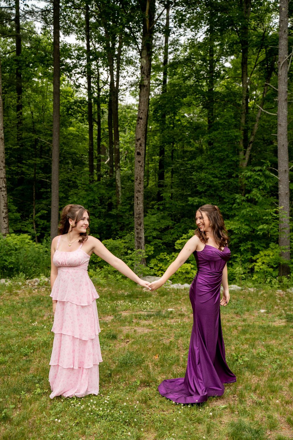 Two women in formal gowns hold hands in a woodland setting, one wearing pink ruffled dress and one in purple.