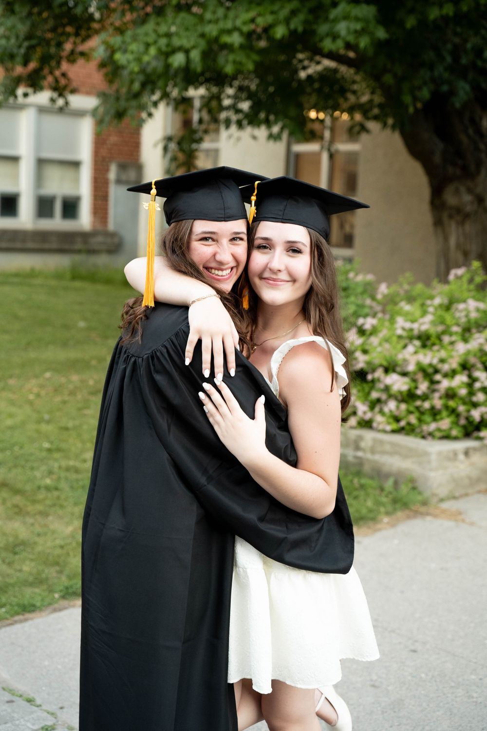 A graduation celebration moment between friends, one in cap and gown, the other in a white dress.