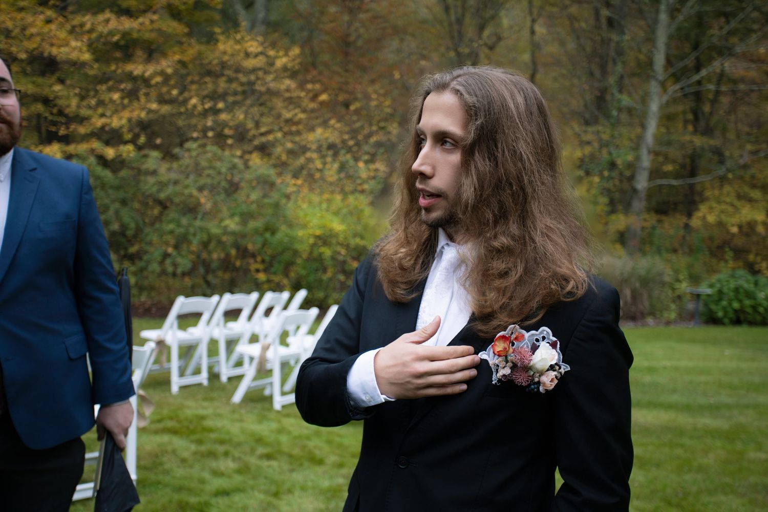 Wedding party gathered outdoors in autumn with white chairs and fall foliage.