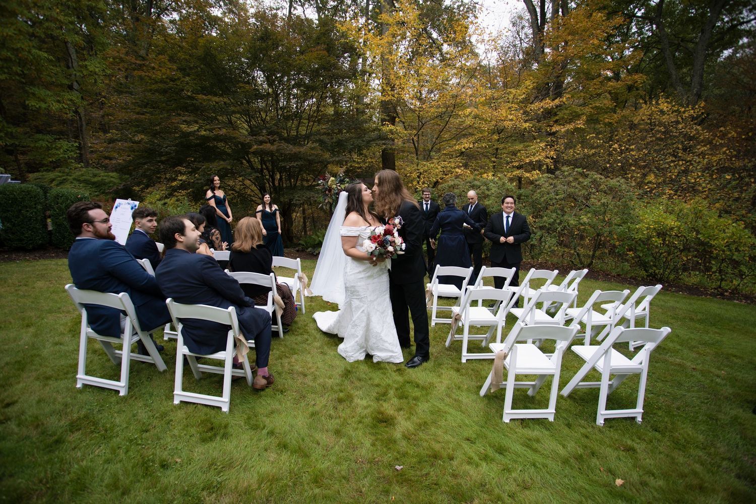 White folding chairs arranged on grass for outdoor wedding ceremony with fall foliage backdrop.