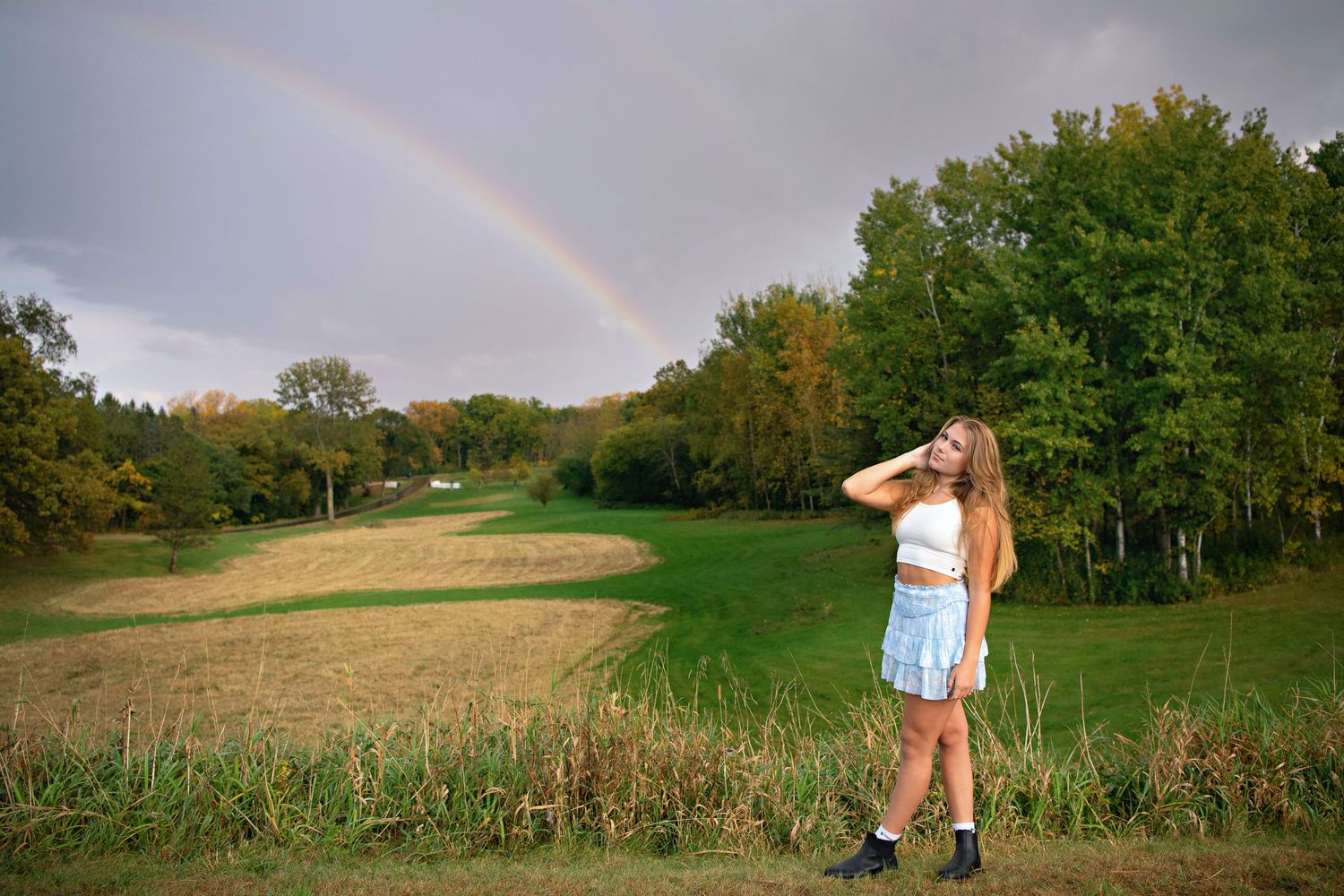 Young woman in foreground with beautiful rainbow arching over a scenic rural field with tall grass and green trees in the background.