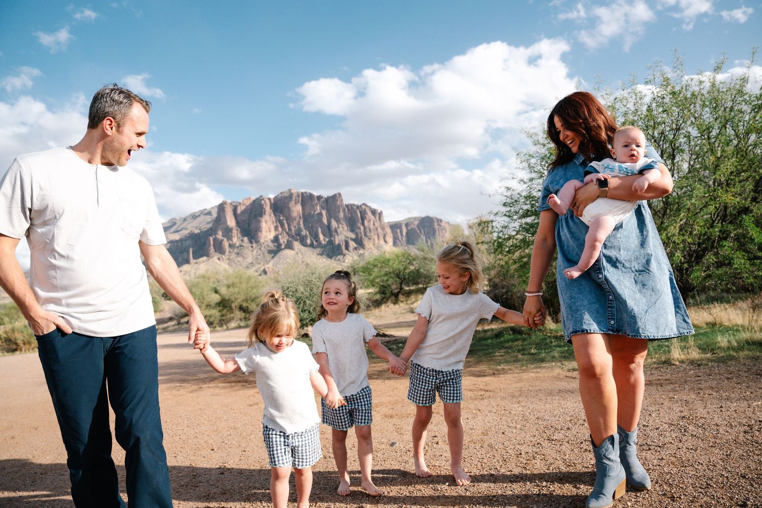 Iowa family photographer – parents and two young boys walking hand-in-hand in desert with mountains, mom carrying baby, joyful family.