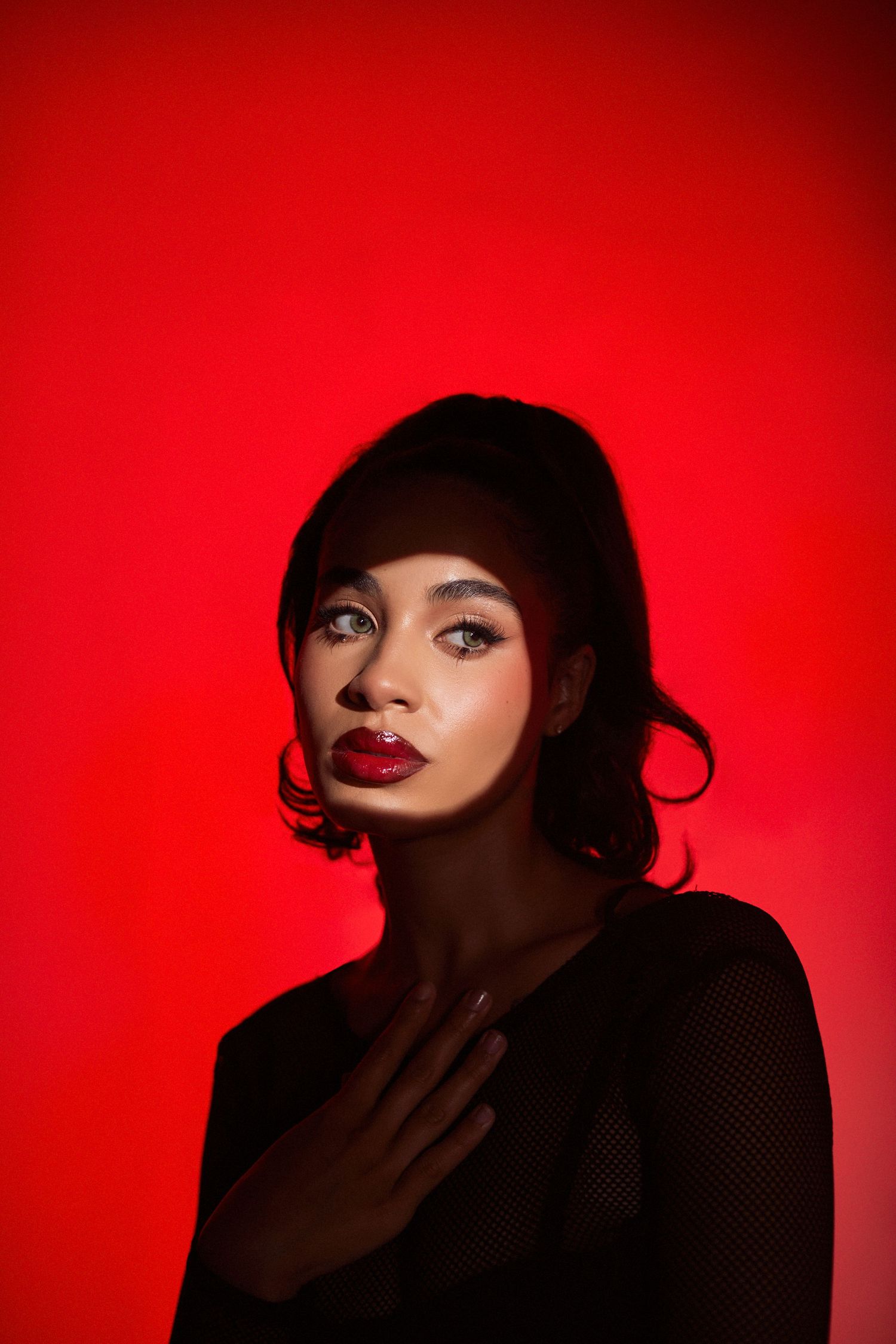 Beauty portrait against red background featuring dramatic makeup and black mesh top.