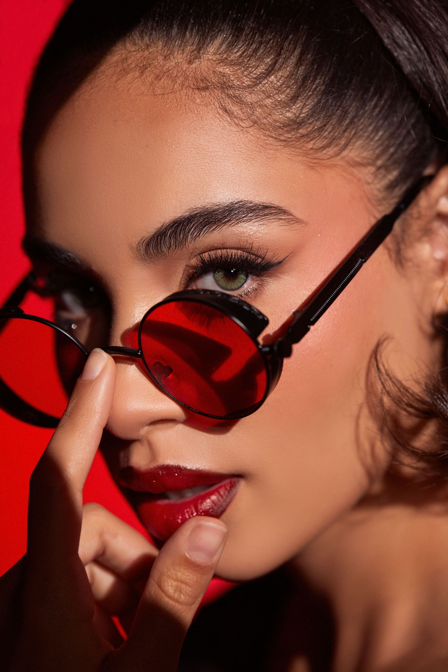 Close-up beauty portrait with red tinted sunglasses and matching lipstick against red background.