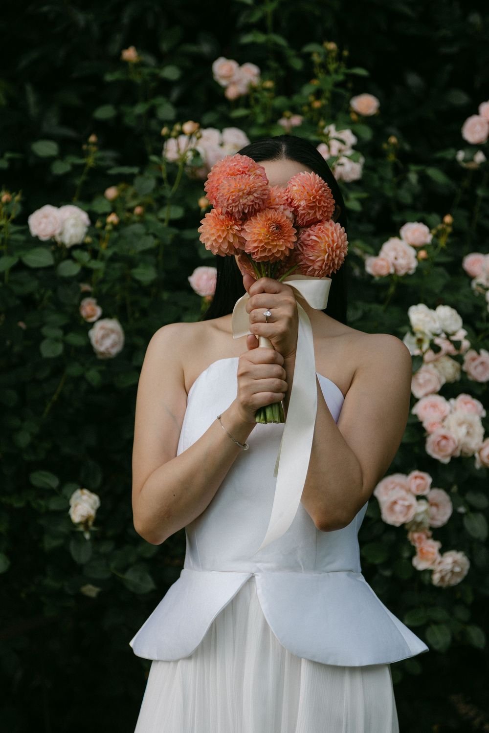 Person in white dress holding coral-colored flowers against a rose garden backdrop.