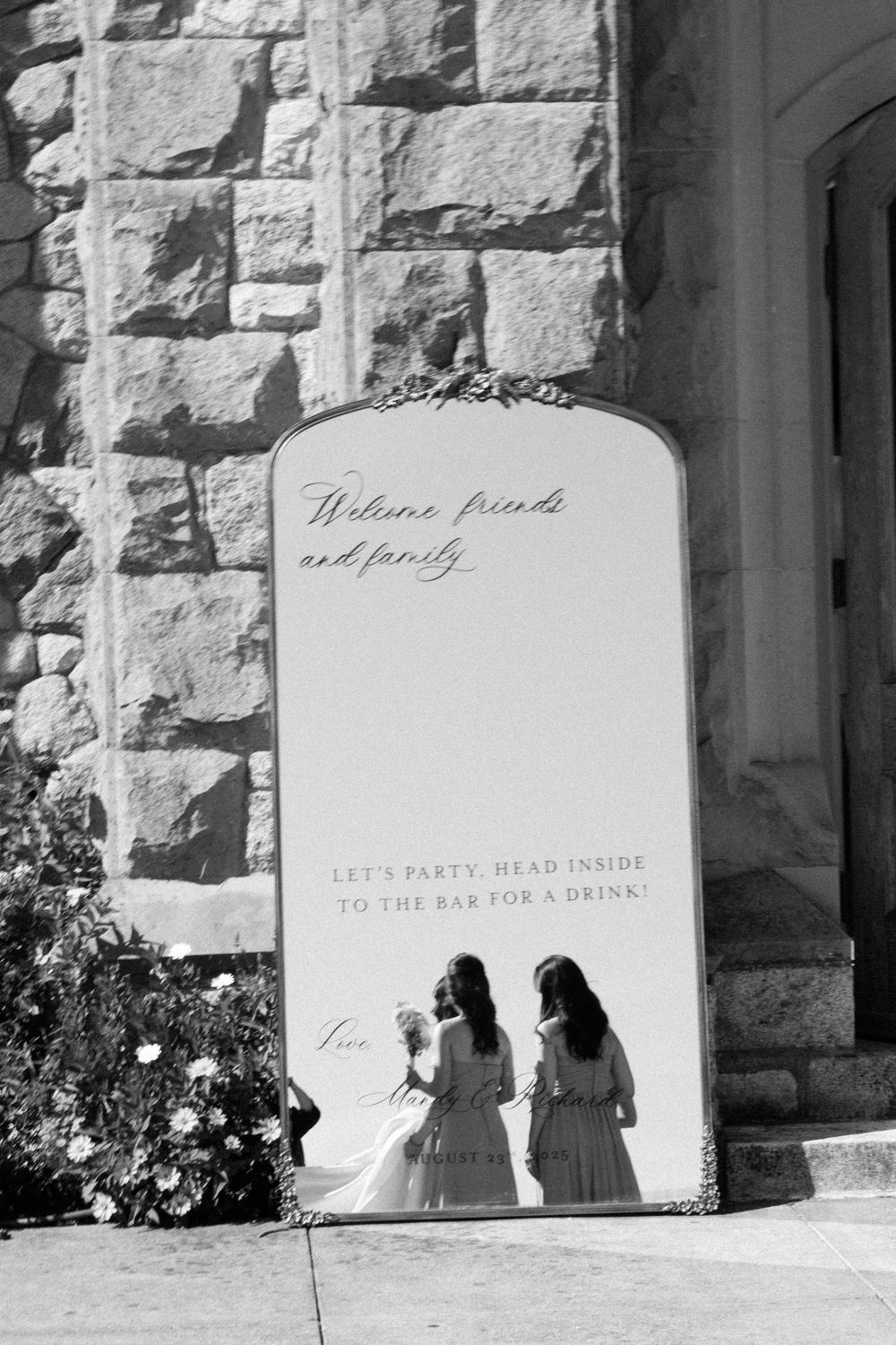 Black and white photo of welcome sign against stone wall with formal event directions.