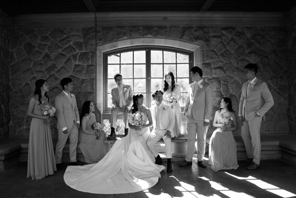 Black and white photo of wedding party gathered in stone room with dramatic window lighting.