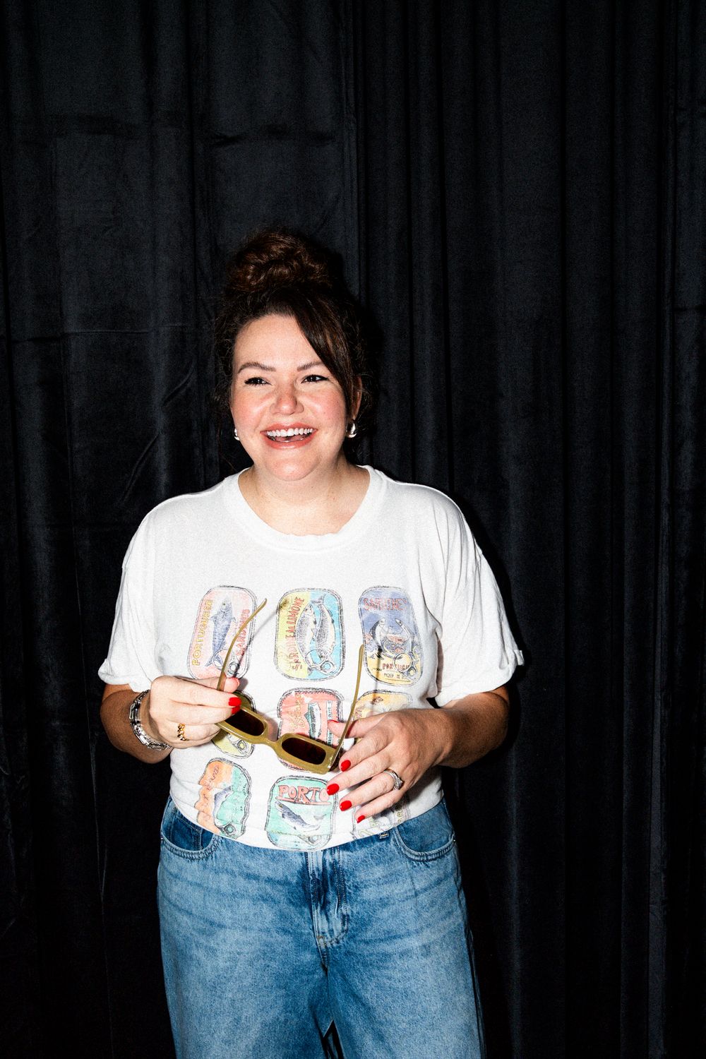 Person in casual white t-shirt and denim poses playfully against dark curtain background.