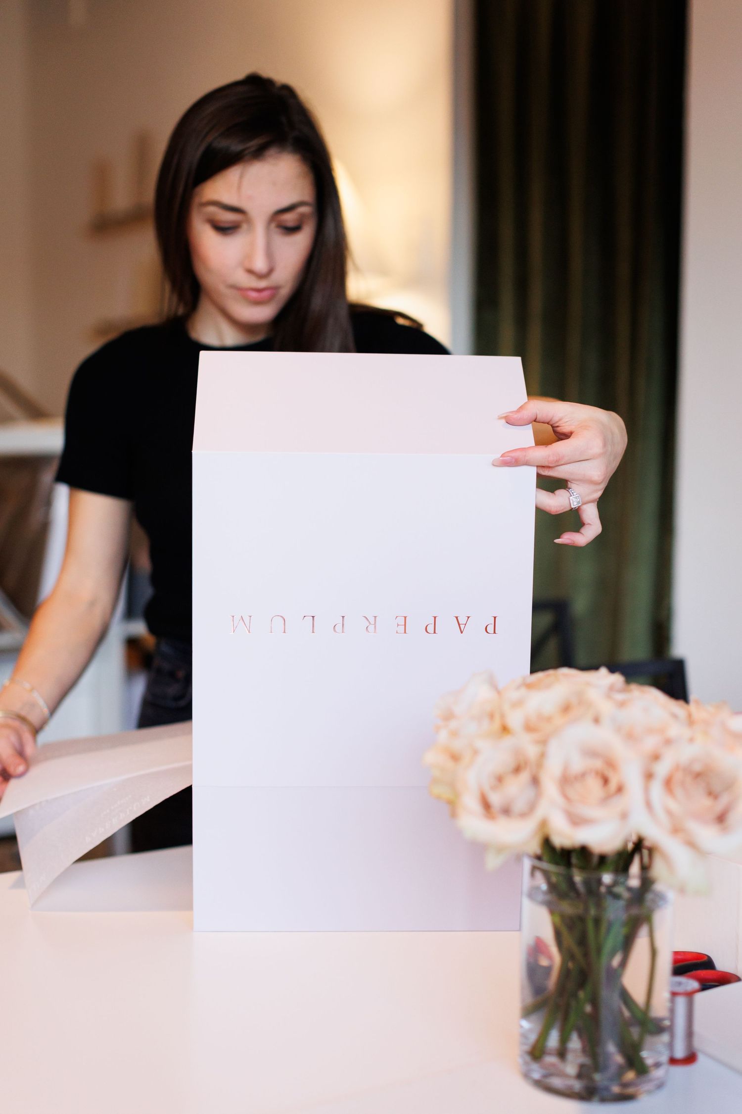Someone wearing black shirt working with paper and documents at desk with pink roses in glass vase.