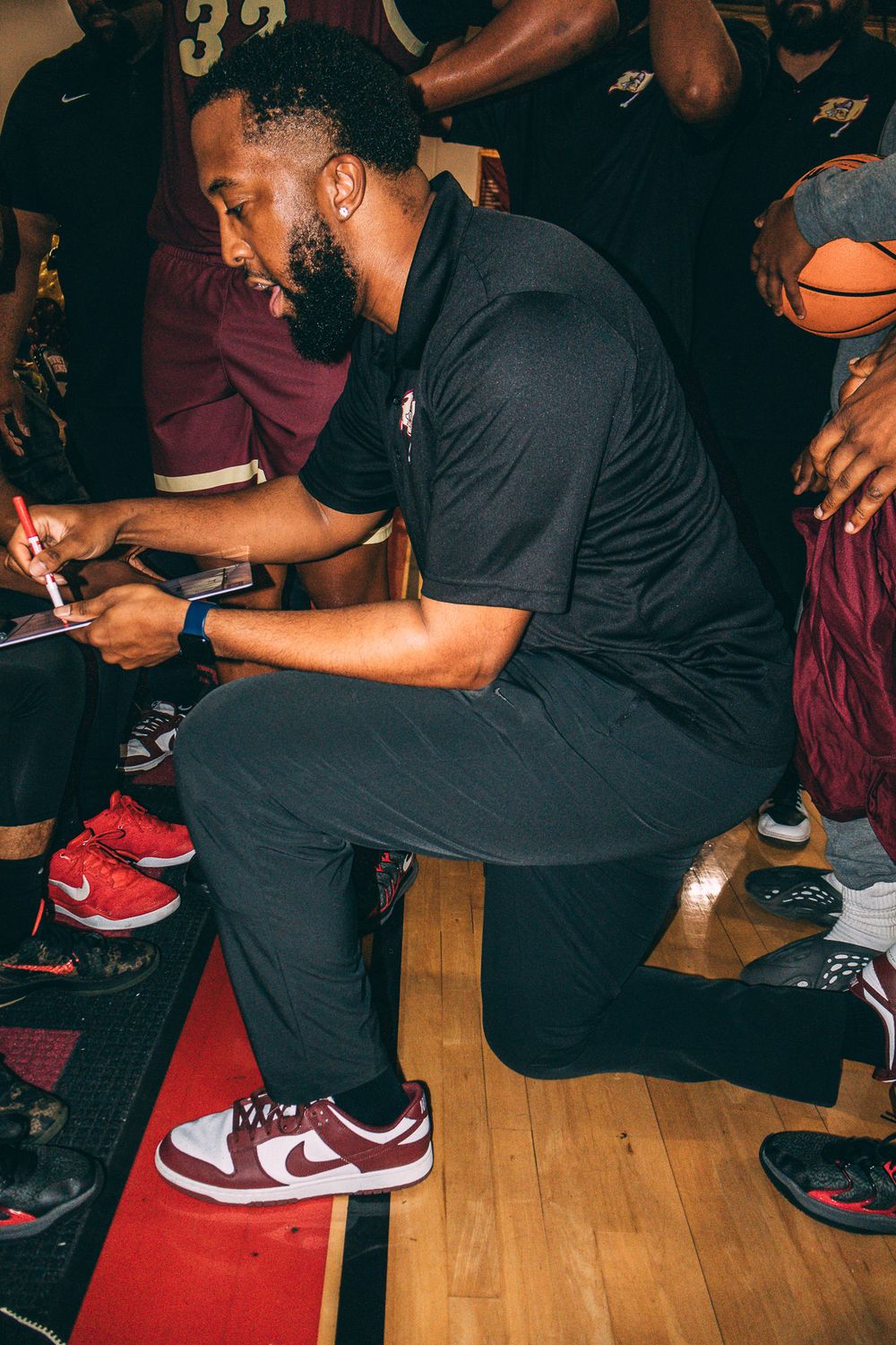 Coach in black attire sitting on sideline bench during basketball game.