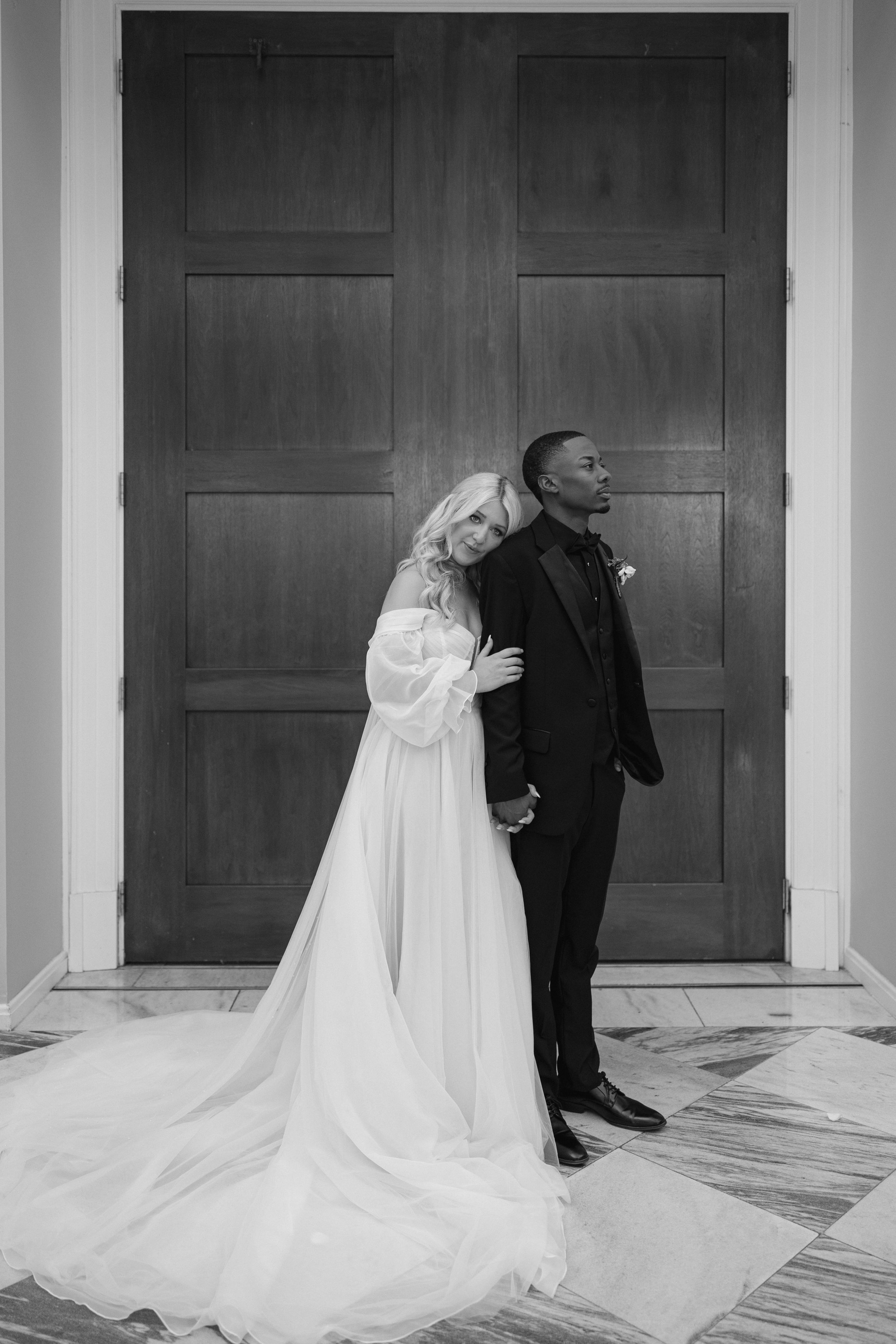 An intimate black and white wedding photograph of a couple standing against wooden doors with flowing white dress details.