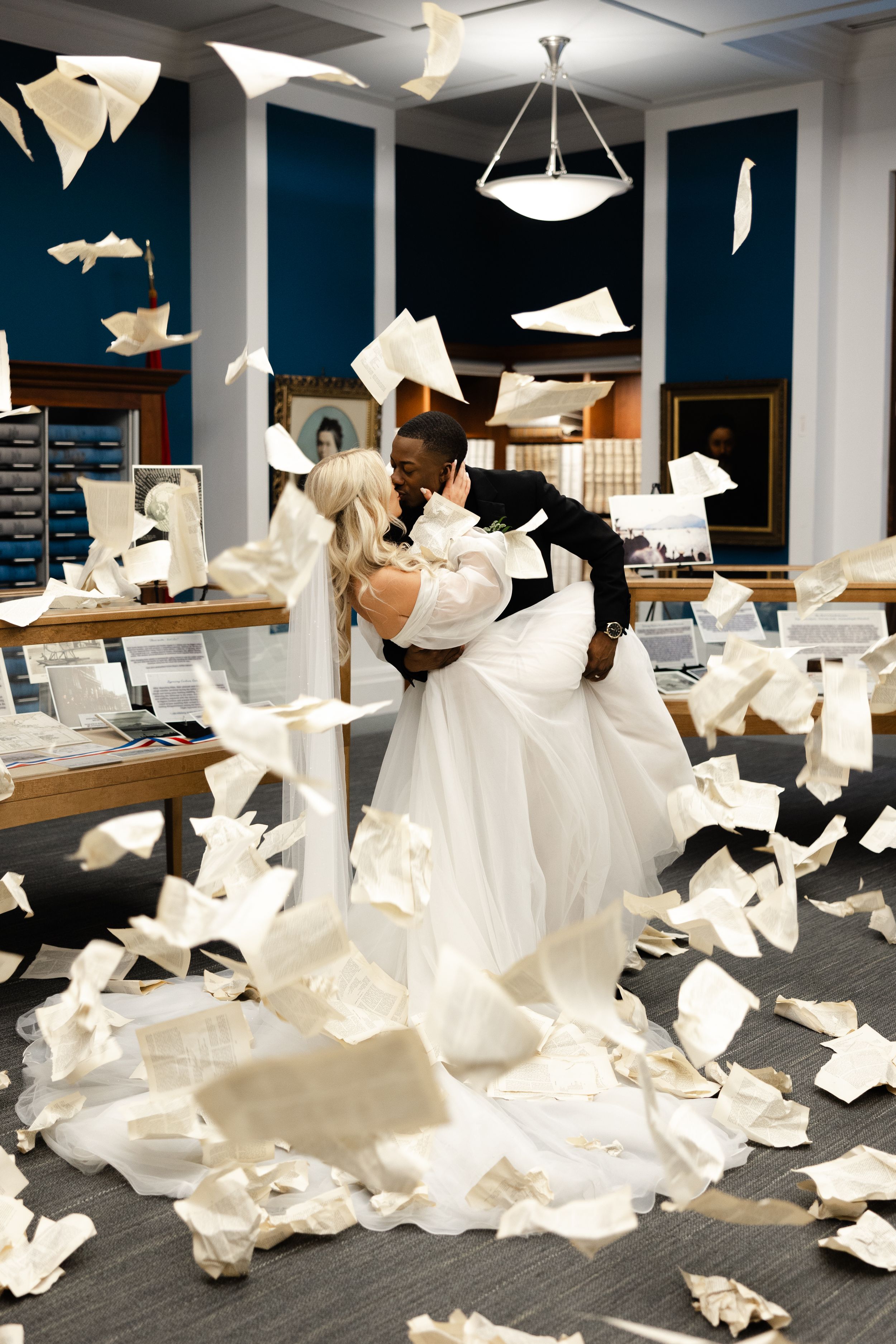 Couple in wedding attire embraces amidst swirling paper and documents scattered across an office floor.