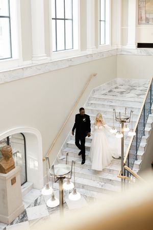 A couple descends an elegant marble staircase in a bright white hall with tall windows and ornate golden railings.