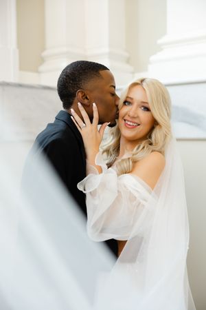 A romantic wedding couple shares an intimate moment in a light-filled space with soft white decor and florals.