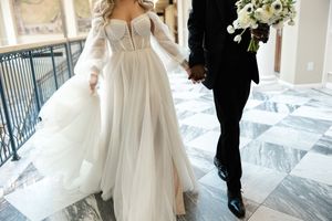 A bride in a flowing tulle wedding gown with sheer sleeves walks alongside a groom in a classic black tuxedo.