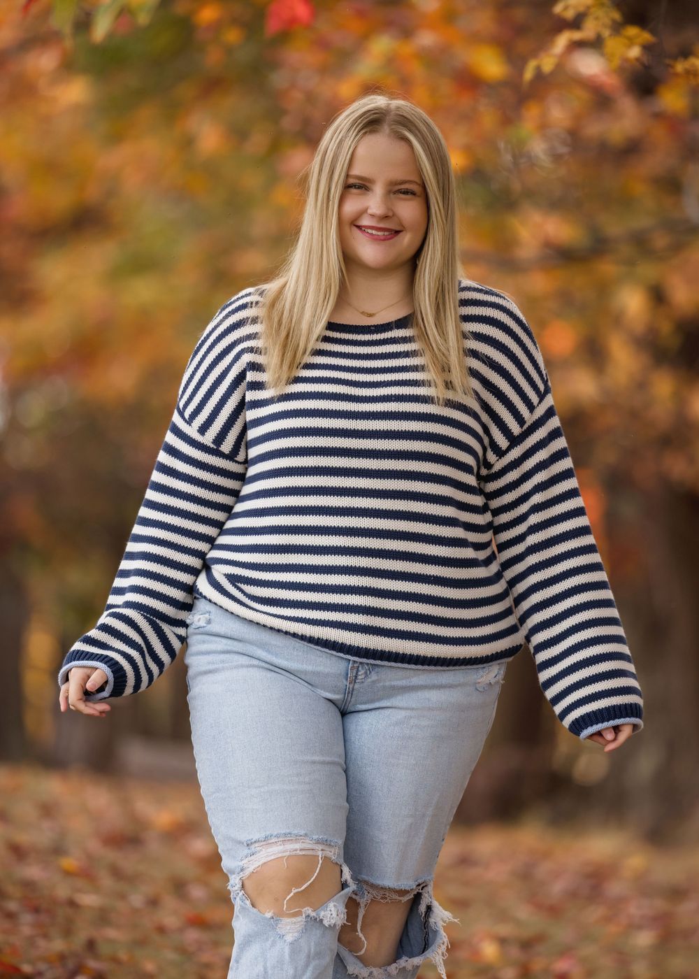 Person in striped shirt and ripped jeans poses playfully against autumn foliage background.