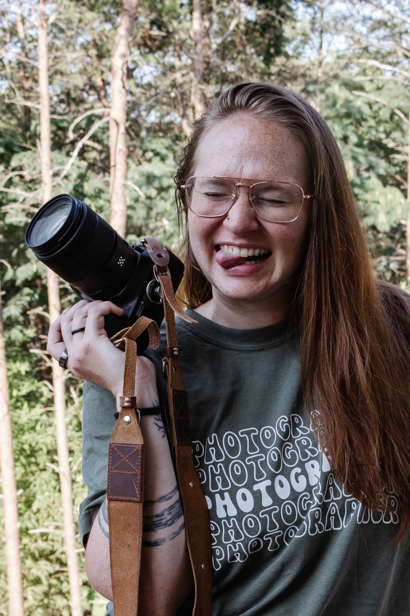 A woman with her eyes scrunched shut smiling while sticking her tongue out holding a canon camera and wearing a photographer shirt