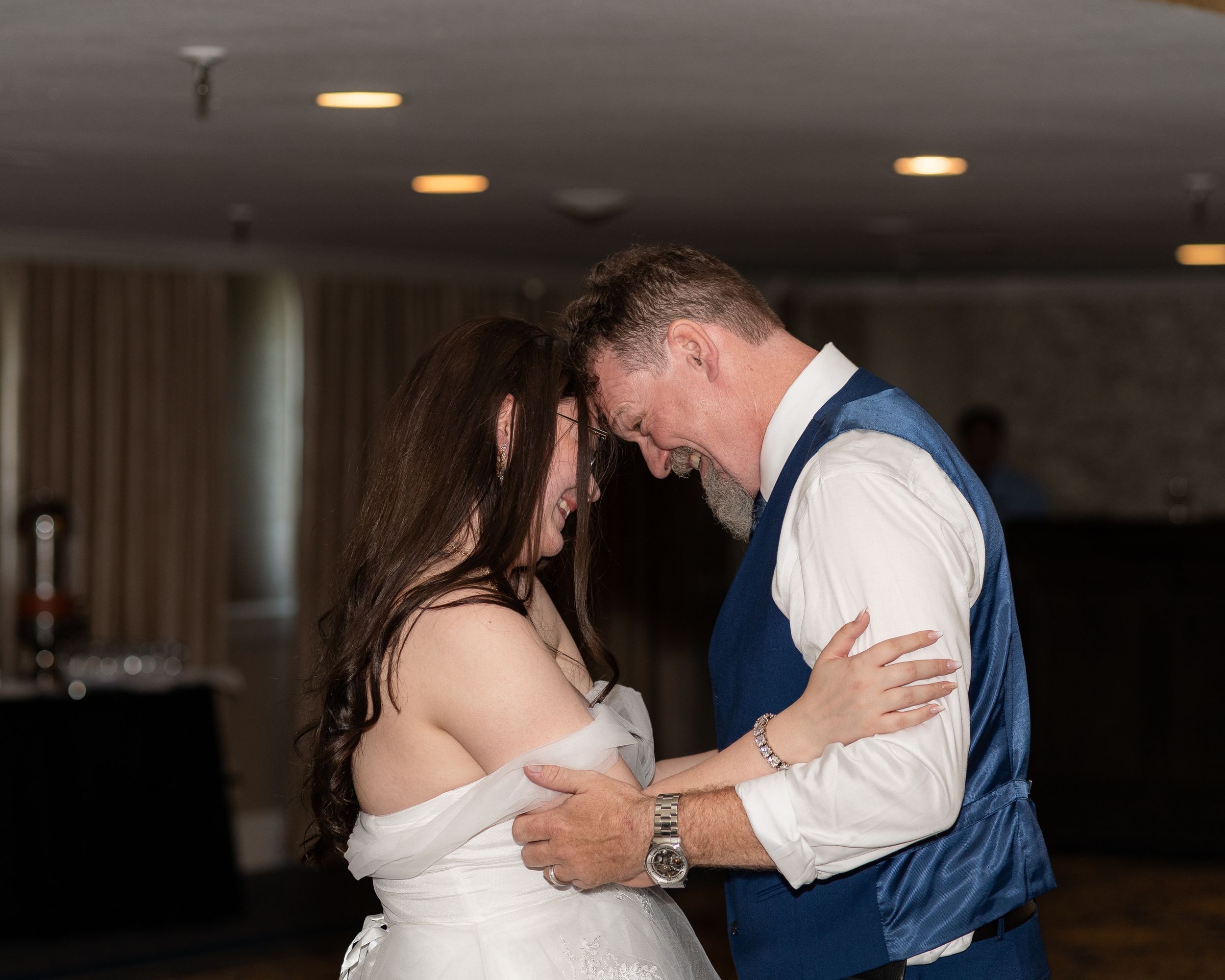 A bride and her father shares an emotional moment while dancing together at an indoor reception venue with soft lighting.