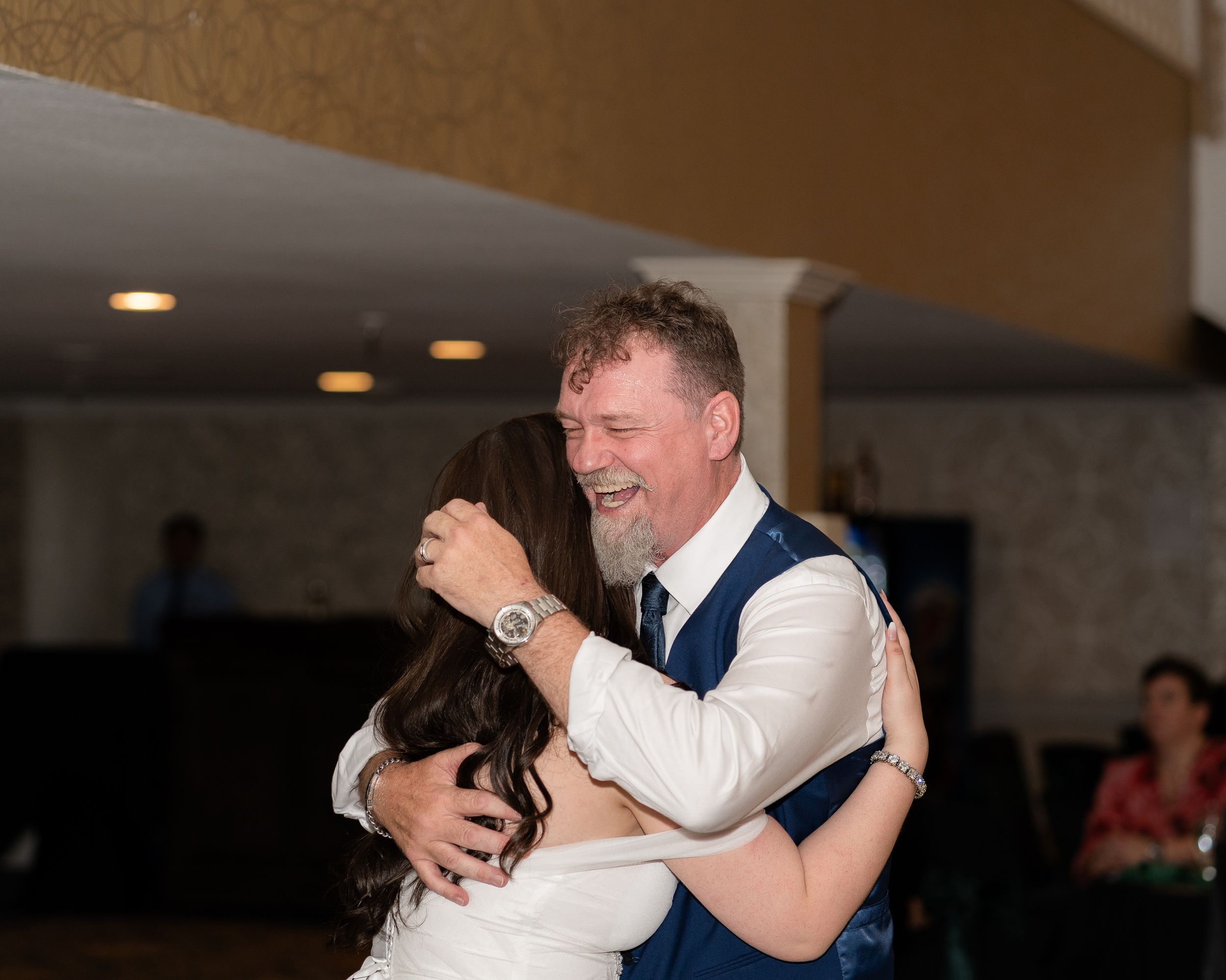 Two people share an emotional embrace while dancing at a wedding reception in a banquet hall.