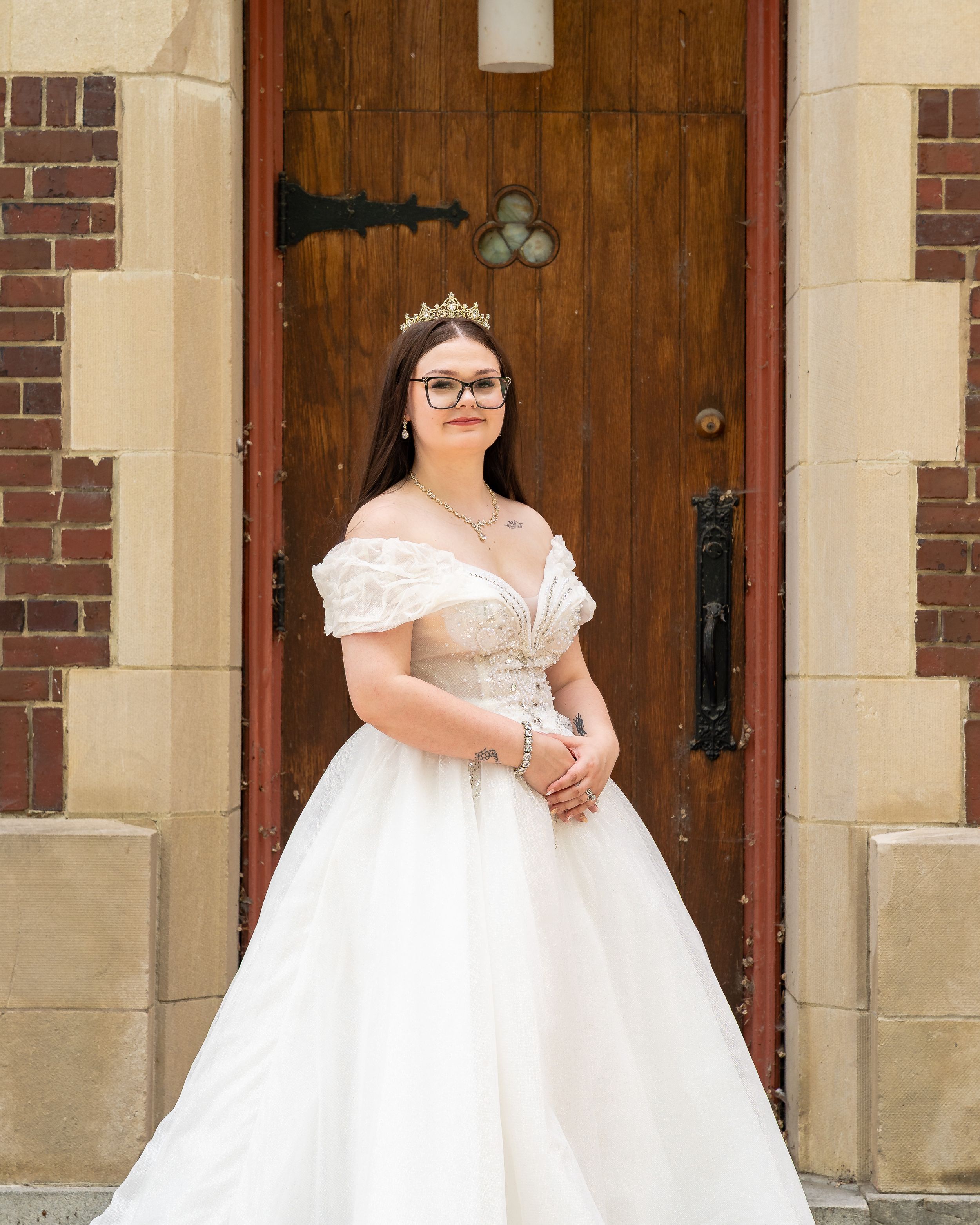 A bride in a white off-shoulder ball gown and tiara stands in front of a wooden church door with brick surroundings.