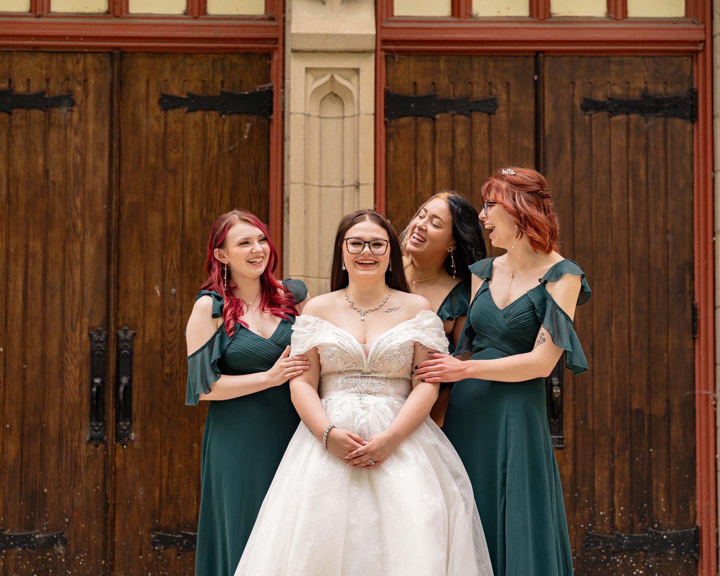 Bridesmaids in emerald green dresses pose with bride in white gown outside wooden church doors.