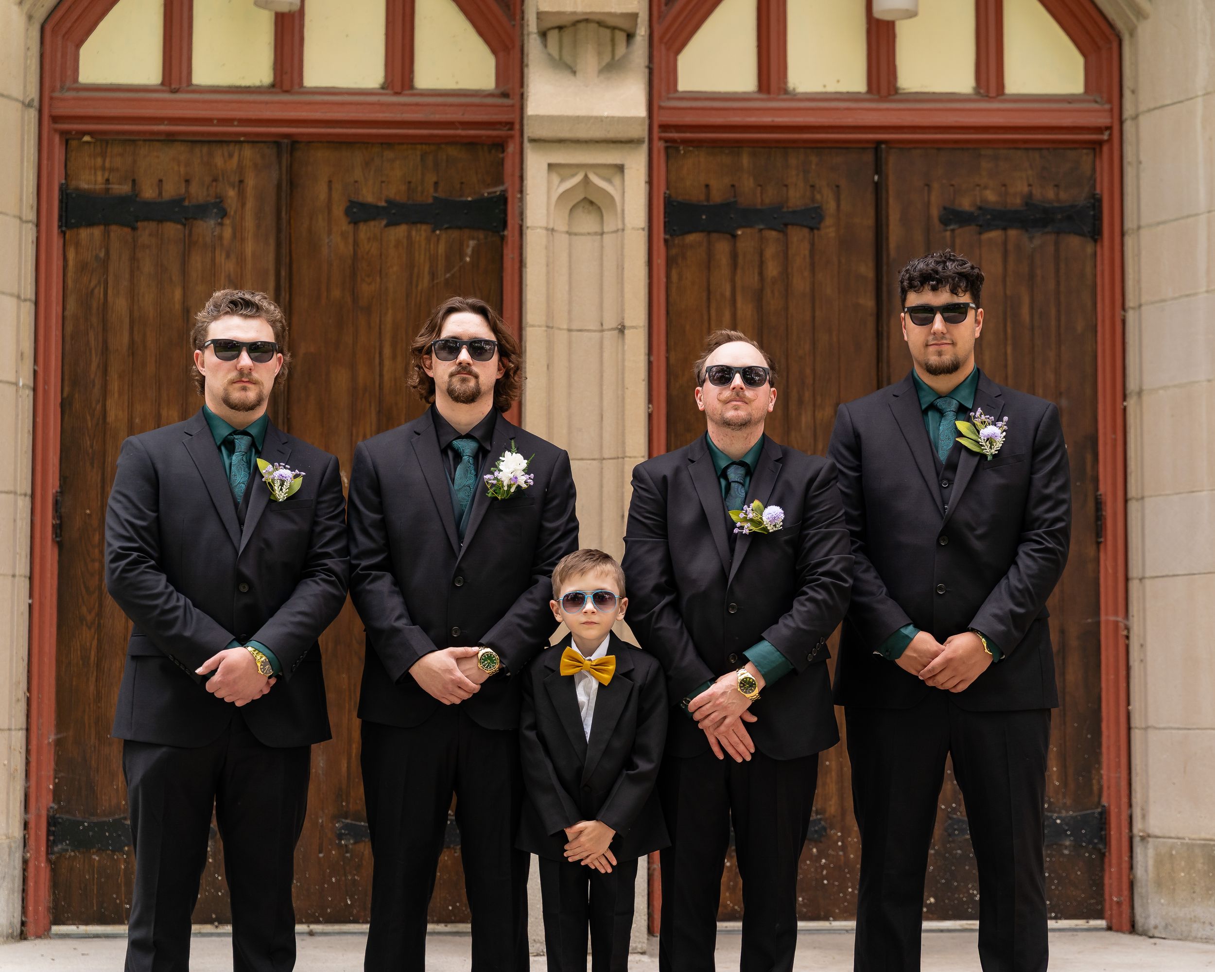 A line of groomsmen wearing black suits and sunglasses pose against a brick wall with boutonnieres.