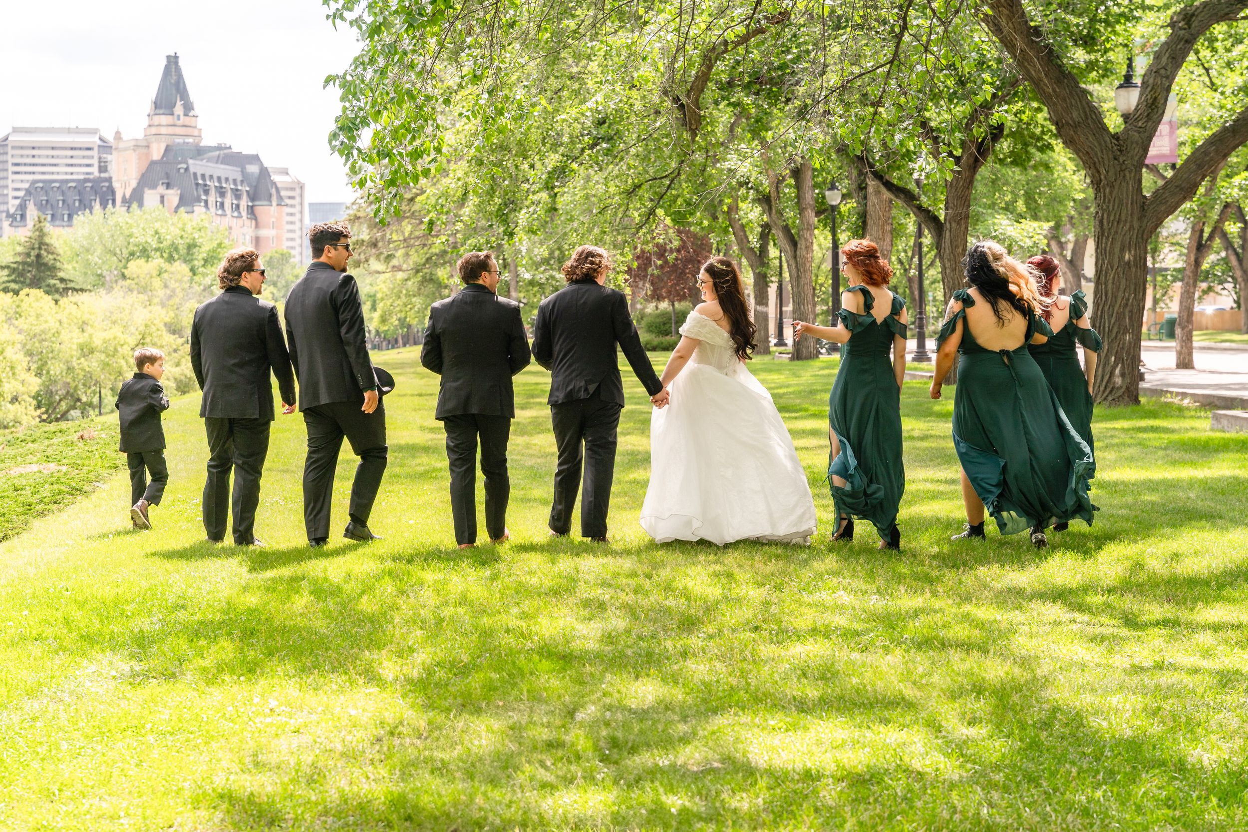 Wedding party walking and laughing together on a green lawn with trees and buildings in the background.