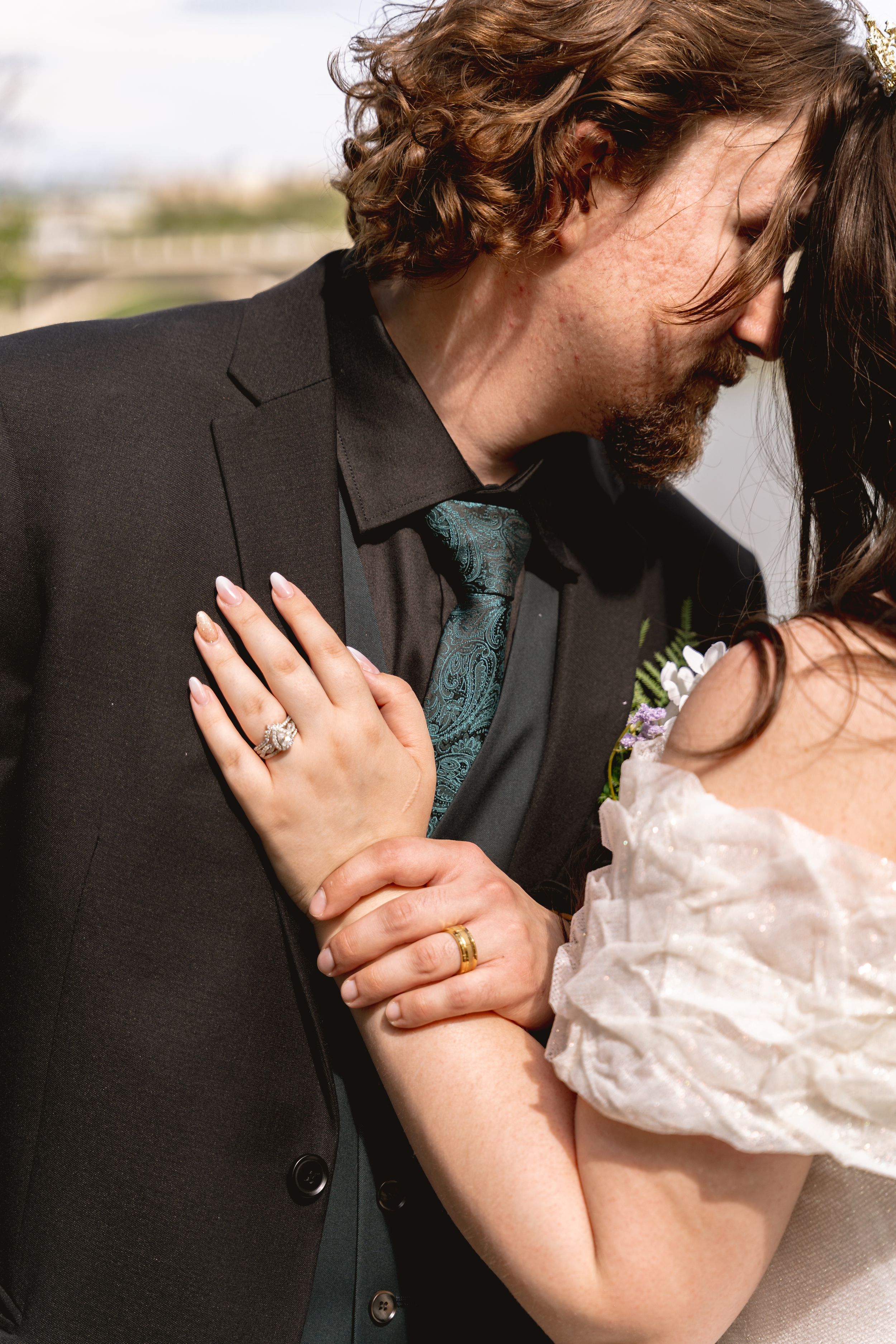 Close up of hands showing wedding rings and formal attire with green tie and white dress in sunlight.