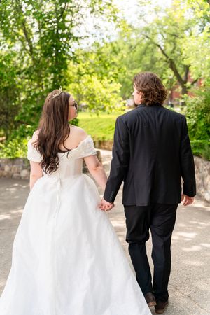A couple walking hand in hand outdoors, bride in white dress and groom in black suit seen from behind.
