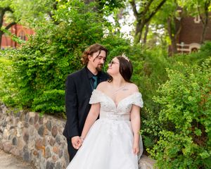 A romantic wedding couple stands together on a stone wall surrounded by lush green foliage in a garden setting.