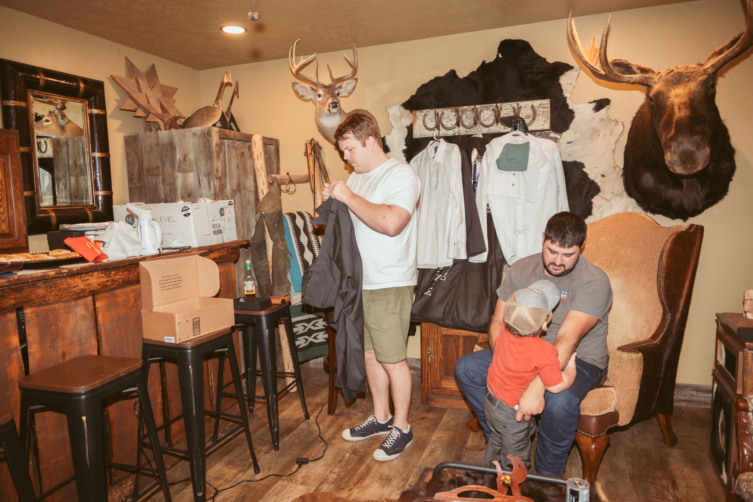 Two people stand in a rustic room decorated with mounted deer antlers and hunting trophies on the wood-paneled walls.