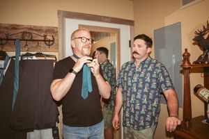 Two people in casual attire stand in a room, with one wearing a black shirt and tie and another in a floral print shirt.