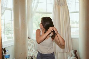 Woman styling her hair in front of a vanity mirror in a sunlit room with white curtains.