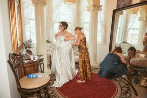 A bridal preparation scene in an elegant room with ornate columns as someone helps adjust a white wedding gown.