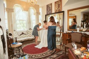 A bridal preparation scene in an elegant Victorian-style room with arched windows and antique furniture on a red oriental rug.