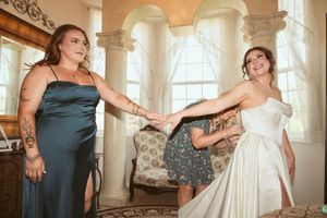 Two people in formal dresses pose playfully together in an elegant room with arched windows and vintage furniture.