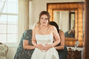 A person in a white wedding dress sits on a green velvet chair in a bright sunlit room with wooden details.