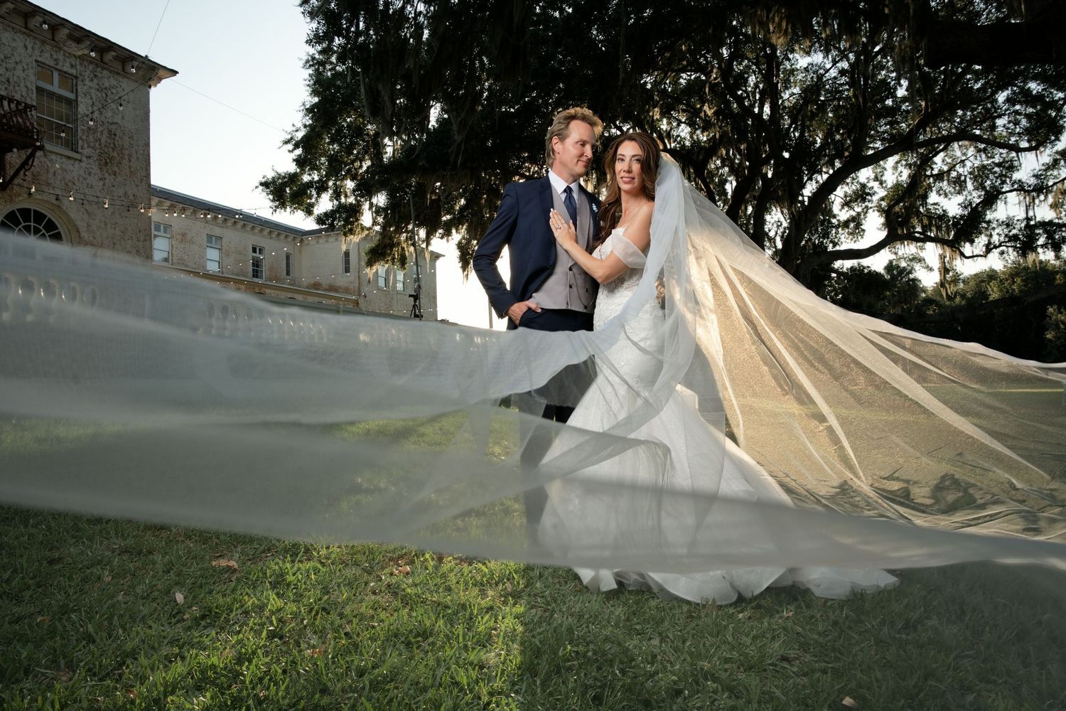 Bride and groom pose outdoors with veil flowing in wind, elegant stone building and oak tree in background. Bella Cosa wedding venue.