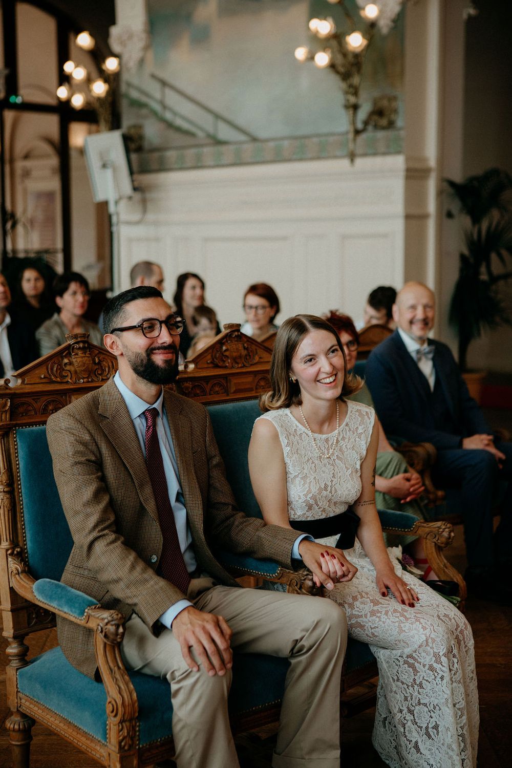 Joyful bride in lace dress and groom in tweed suit laughing together during wedding ceremony in grand hall.
