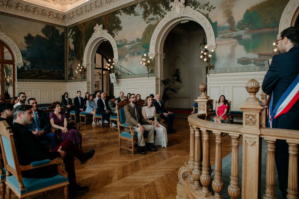 Elegant French civil wedding ceremony in ornate hall with murals, guests seated in blue chairs facing officiant at podium.